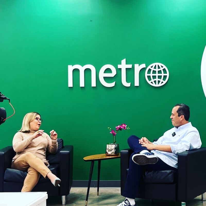 Frente a una pared verde con el logo del periódico Metro en blanco, la periodista y editora Aiola Virella aparece sentada, entrevistando al también periodista Rafael Lenín López, que la observa, también sentado.