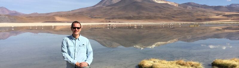 EL investigador Adam Charles Simon aparece con manga larga, gafas oscuras y sosteniéndose las manos frente a un paisaje que incluye un lago y un volcán.