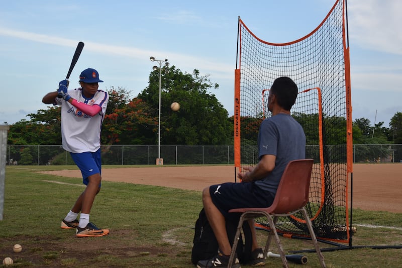 Eddie Martínez Medina batea pelotas frente a una malla de práctica. Lo acompaña su padre, Eddie Martínez Ponce.