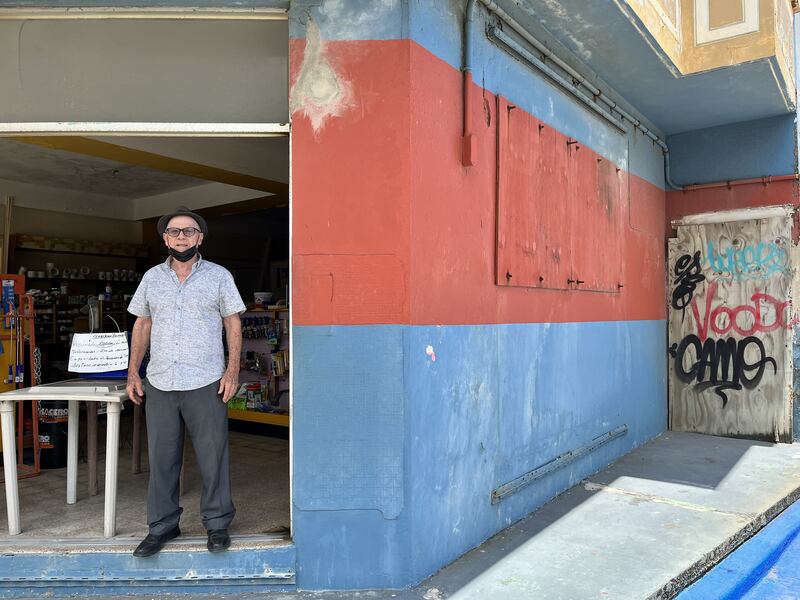 Don Luis Benabe de pie en la entrada de su ferretería, un edificio color rojo y azul con una pared con grafitis.