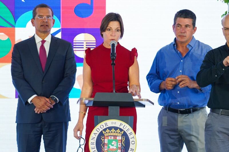 Mariela Vallines Fernández al centro, hablando desde un podio con el sello del Gobierno de Puerto Rico. A su izquierda está el gobernador Pierluisi y a su derecha Jacobo Ortiz Blanes.