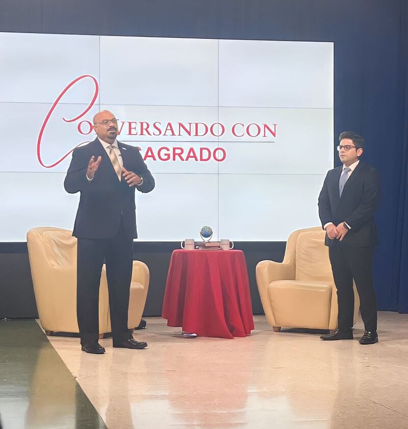 En una toma lejana, Rolando López y Francisco Parés están de pie, frente a dos sillas amarillas y una pantalla que lee "Conversando con Sagrado".